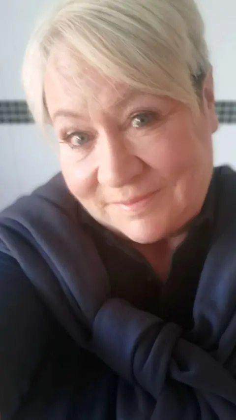 A photo of Kelly Faiers. She has blonde hair and is smiling at the camera. She is wearing a dark coloured top and has a dark blue sweater tied around her neck.
