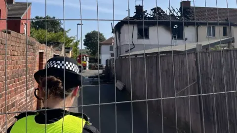 Louise Hobson/BBC A police officer is stood facing a badly burnt property. The roof has caved in and exposed the wooden beams. An alley runs down next to the property and it has a wooden fence on one side