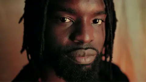 A close-up of Meji Alabi, with beard and dreads, looking into the camera