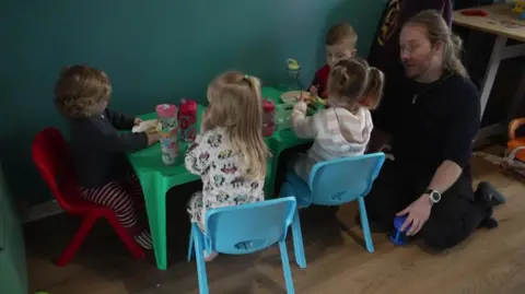 Ash Crouch kneeling beside a green plastic table with four toddlers sitting eating