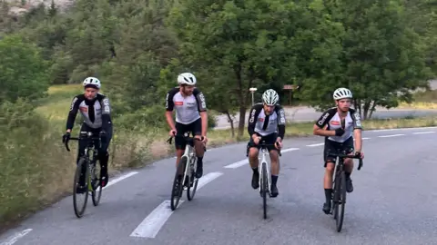 Michael Mansell Four cyclists riding up a hill in the Alps, wearing red and white jackets and black shorts or trousers.
