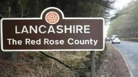 Brown road sign which reads 'Lancashire The Red Rose County' in white letters on the side of a road