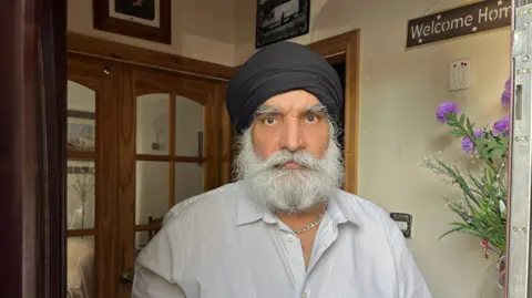 A man in a black turban and white shirt with very thin stripes on it looks at the camera with a solemn face. He has a grey, bushy beard and moustache. He stands in the doorway to his home.