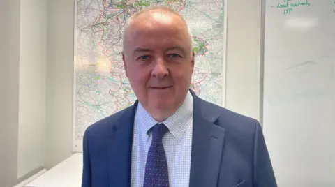 A man with very short grey hair is wearing a dark blue suit jacket with a dark blue patterned tie, and white shirt with light blue squares on it. He stands in front of a whiteboard and a map of Staffordshire.