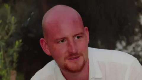 Matthew Charnock smiles and looks just to the right of the camera, wearing a white shirt, with trees or shrubs seen blurred in the background. He is bald and has a ginger beard.