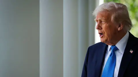 US President Donald Trump walks out of the Oval Office for a signing ceremony for the Take It Down Act, in the Rose Garden of the White House in Washington DC on May 19. He wears a blue suit and tie and appears to be saying something. 