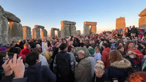 A large crowd of people gathered around Stonehenge as the sun rises over the horizon. The sky is light blue and one the left side of the stone sculptures are golden from the sunrise.