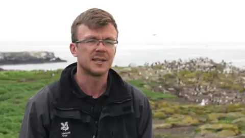 Tom Hendry standing on Inner Farne with birds on cliff edge and North Sea in background. He has short brown hair and wears glasses