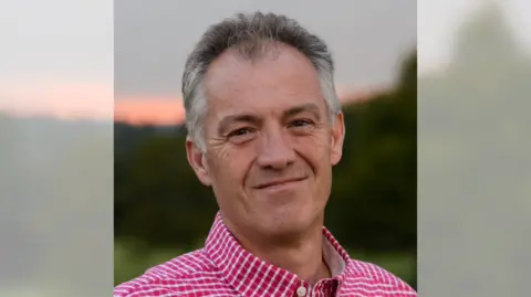 Acorn Bioenergy A man with a red and white check shirt smiling at the camera in front of a blurred sunset