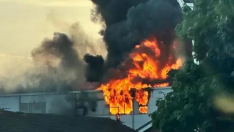 Large plumes of thick black smoke coming from the roof of a building where big orange flames are climbing out 