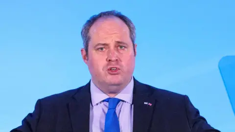 Reuters Alex Wilson is seen from the chest up, wearing a dark suit with purple shirt and blue tie. He is in the middle of giving a speech against a blue background