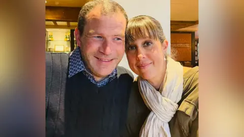 Handout A man with greying hair, and a black jacket, is sitting next to a woman with dark blonde hair, a white scarf and khaki jacket.