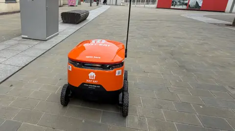 The orange coloured robot, with Just Eat branding in white, is travelling towards the city's Keel Square. It has six chunky rubber wheels and a large pole which carries a flashing orange flag to indicate it is near. In the distance is the under-construction Culture House. 