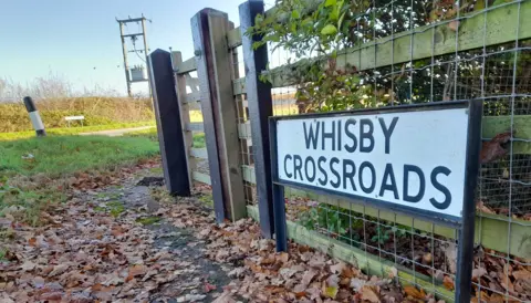 LDRS A sign on a fence which reads "Whisby Crossroads". The road is in the background and there are leaves along a path