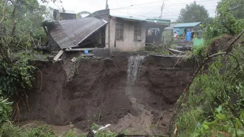 CHARISM SAYAT/AFP via Getty Image A man in a blue rain poncho looks at a landslide-hit residential area surrounded by greenery and trees in Guinobatan town, Albay province, south of Manila. A small concrete house, with a tin roof collapsed beside it, sits on the edge of a precipice that has been stripped to dirt by a landslide, with water running off it.