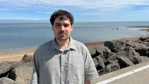 A young man with dark hair and stubble stands on a coastal path in front of a pile of grey rocks, with a shingle beach and the sea behind him.