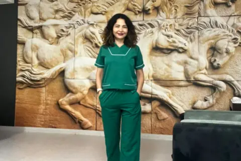 Firdevs in green medical scrubs stands indoors in front of a large sculpted wall mural depicting galloping horses in relief.