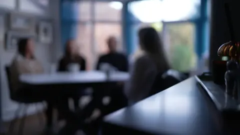 A blurred image to protect the identity of Sarah, showing BBC correspondent Judith Moritz, Sarah and her parents, sitting together at a table next to a large window