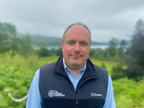 Outward Bound chief executive Martin Davidson, a grey-haired man with a sun-kissed complexion, a blue shirt and a navy, branded Outward Bound fleece, stands in the greenery with a stunning loch view behind him.