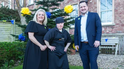 Andy Barnham Three people standing outside a hotel in Minehead. The woman on the left has blonde hair and is wearing a black dress. The young woman in the middle is wearing a black t-shirt and hat. The man on the right is in a navy suit. A building, a hedge and a bench are in the background.