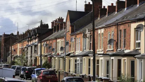 Typical terrace houses in a residential block with large front windows jutted out and cars outside. 
The houses are red brick with yellowy cream paint on the downstairs window frames.