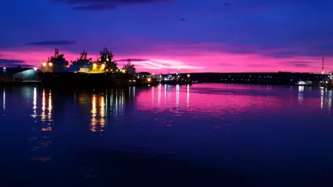 Ian Jamieson A harbour at night with a dark blue and pink sky and the lights from a number of boats shining out and reflected in the water