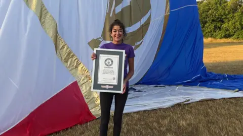 Jo Bailey, wearing black leggings and a purple t-shirt, standing in a field in front of a large deflating hot air balloon, which is gold, white, red and blue. She is holding a framed award.