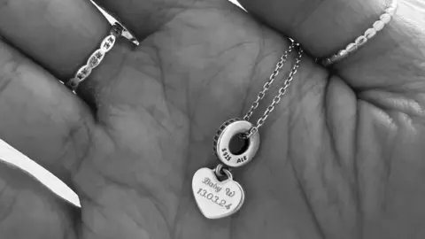 A black and white image of the palm of a hand holding a necklace with the words Baby W 13.03.24