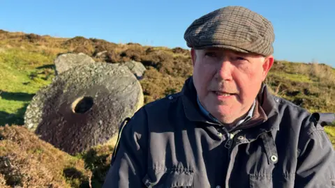 A man sitting outside with a mill stone in the background behind him. He is wearing a navy jacket and a cap.