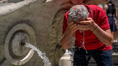 A man cools himself at a water fountain. He is wearing a red T-shirt and blue shorts and has both hands over his face, from which water is dripping towards the ground.