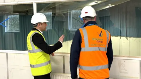 Wiltshire College Two men in high-vis vests are standing with their backs to the photographer and looking through a window. They are both wearing white hard hats.