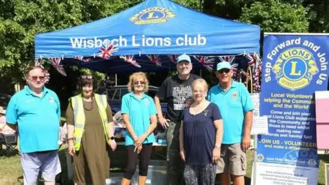 WISBECH LIONS Three men and three women on a sunny day smile at the camera as they are photographed outside. They are stood in front of a small gazebo with Wisbech Lions written on it a banners beside it.