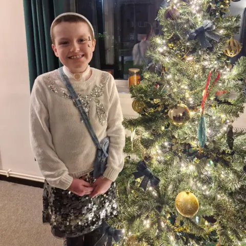 Family A girl is standing next to a Christmas tree. She is wearing a silver sequinned skirt and a cream jumper with sequins embroidered on it in a bow pattern. She has very short brown hair and is wearing a silver headband.