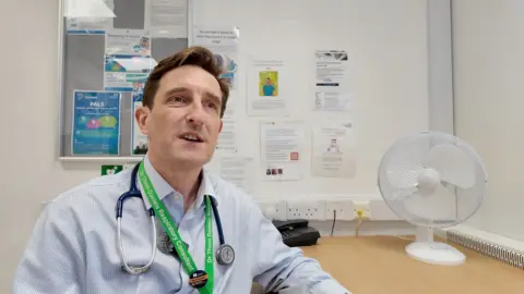 A doctor wearing a light blue shirt and with a stethoscope around his neck sits at a desk.