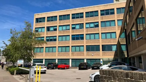 Local Democracy Reporting Service A sand-coloured five-storey office block showing small windows grouped in threes on each floor and a flat roof. There are cars parked in front of the building. A tree is to the left of the building. There is also a car park barrier to the left.