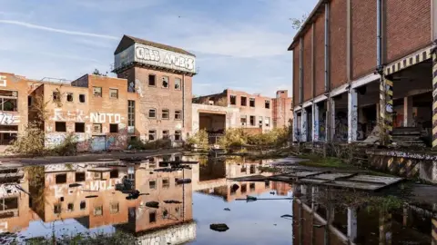 Old industrial brick buildings. The land between two separate blocks is muddy and water covers the floor, reflecting the buildings in it.