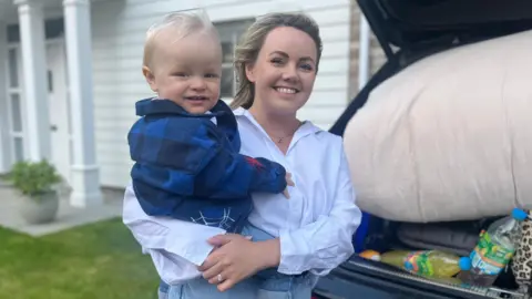 Catherine Fleming and her 16-month-old son Luca standing at the back of an open car, smiling at the camera