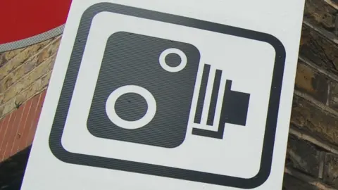 Black and white traffic camera logo against a brick wall