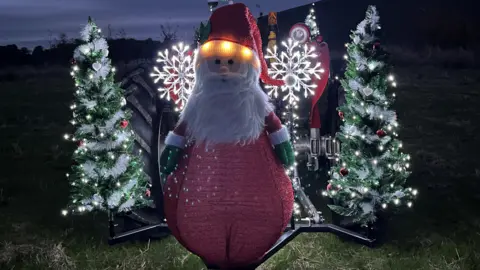 Brian Chandler A model of a Father Christmas sits at the front of a tractor and Christmas trees are attached to either side. It is lit with fairy lights.