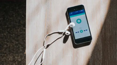 Getty Images An iPhone is sitting on a bare wooden table, with a pair of white wired earphones lying beside it. A shadow is edging into the image from the right. On the screen of the phone a music app is open.
