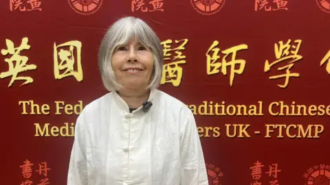 The picture features a woman with white hair wearing white traditional Chinese clothing. Behind her is a wall with large Chinese font, with an English translation below. The wall features Chinese symbols.