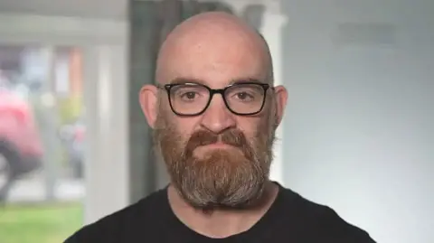 Keith Macaldowie, a bald man with a brown/red/grey beard looks sternly into the camera. He wears glasses and a black t-shirt.