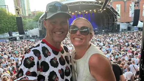 Kaela and Jonathan Rigg stand together at a concert. She is wearing sunglasses and he is wearing a bucket hat. There is a large crowd and a stage in the background. 