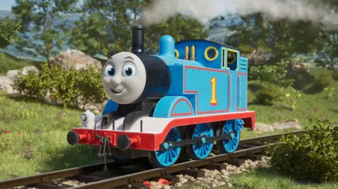 PA Media A blue train engine with a grey smiling face. It has a yellow number one on the side of it, and the track it is running along is surrounded by countryside.