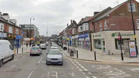 A busy high street lined with different types of shops. The centre of the road is full of traffic.