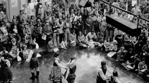 Mind Body Spirit Festival Crowds sit around a stone dance floor as people dressed in long white shirts and skirts dance together. 