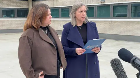 Laura Foster/BBC Sonia Martin-Coppen and Kelly Fretwell stand outside the concrete Chelmsford police station building. Sonia is brunette wearing a brown coat. Kelly is in a blue coat and has blonde hair. There are microphones in front of them.
