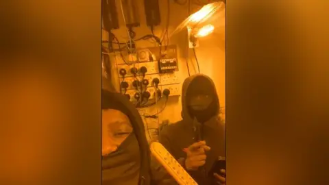 Nottinghamshire Police Mobile phone image of two men in balaclavas, one holding a knife, in a room lit with very yellow light, in front of a series of wall mounted electrical sockets