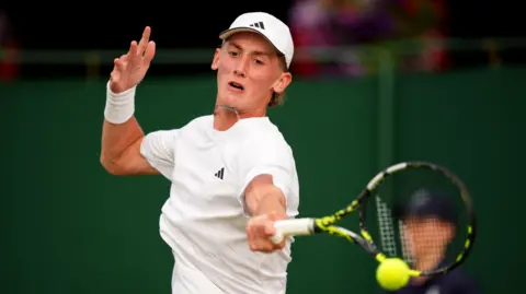 Henry Searle, a young man with blond hair, is pictured while playing tennis. He wears a white shirt and cap and is holding the racquet in his left hand while hitting the ball.