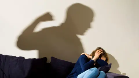 Dominic Lipinski/PA Wire A shadow of a man with a clenched fist as a woman cowers in the corner of a room, on a sofa. 